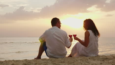 Ein-Junges-Paar-Trinkt-Wein-Am-Meer-Bei-Sonnenuntergang-Sitzen-Sie-Auf-Dem-Sand-Klirren-Gläser-Jahrestag