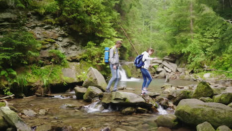 Ein-Junges-Touristenpaar-Mit-Rucksäcken-überquert-Einen-Bergfluss-Im-Wald-HD-Video