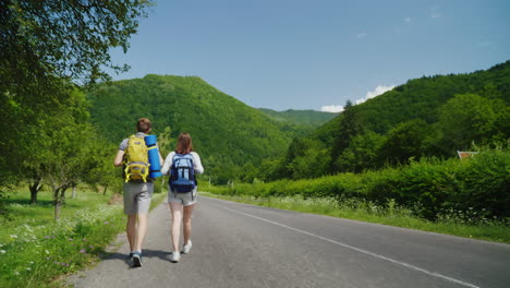 A-Couple-Of-Tourists-With-Backpacks-Are-Walking-Along-The-Asphalt-Road-Towards-The-Beautiful-Green-M
