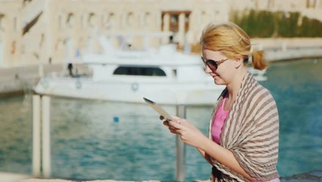 A-Female-Tourist-Enjoys-The-Tablet-Against-The-Backdrop-Of-The-Bay-With-Yachts