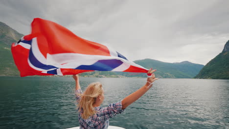 Eine-Aktive-Frau-Mit-Einer-Großen-Norwegischen-Flagge-Steht-Am-Bug-Eines-Kreuzfahrtschiffes-Fjordkreuzfahrt-In