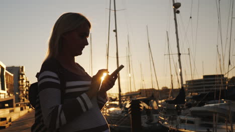 Silhouette-Einer-Frau-Mit-Einem-Smartphone-In-Der-Nähe-Des-Piers,-An-Dem-Viele-Yachten-Vor-Anker-Liegen