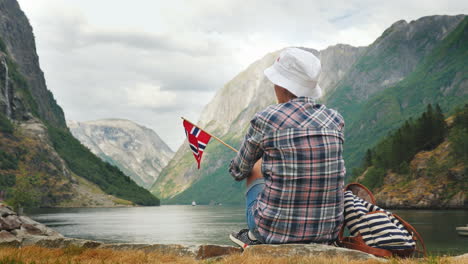 A-Woman-With-The-Flag-Of-Norway-In-Her-Hand-Admires-The-Beautiful-Fjord-Tourism-In-Scandinavia-Conce