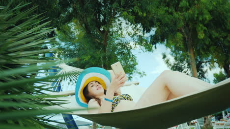 A-Modern-Young-Woman-In-A-Wide-Brimmed-Hat-And-Open-Swimsuit-Enjoys-A-Holiday-On-A-Hammock-Reads-New