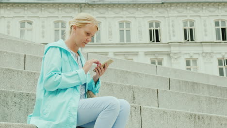 A-Woman-Uses-A-Smartphone-Sits-On-The-Steps-Against-The-Background-Of-A-Beautiful-Building-A-Trip-Th