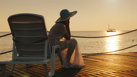 A-Woman-Looks-At-The-Sunrise-Over-The-Sea-Sits-On-A-Sunbed-In-The-Distance-A-Fishing-Boat-Is-Visible