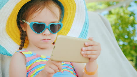 Am-Strand-Mit-Einem-Smartphone-Kleines-Mädchen-Spielt-Auf-Dem-Smartphone-Sitzt-Im-Großen-Hut-In-Der-Chaiselongue