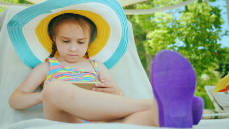 A-Cute-Little-Girl-In-A-Large-Multi-Colored-Hat-In-A-Bathing-Suit-Is-Sitting-On-A-Lounger-And-Playin