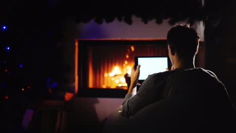 A-Man-Is-Resting-By-The-Fireplace-Decorated-For-Christmas-Using-A-Tablet-Ordering-Holiday-Gifts