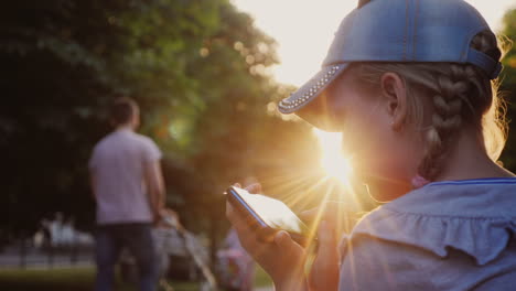 A-Child-Plays-On-A-Smartphone-Sits-In-A-Park-On-A-Bench