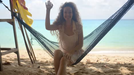 Smiling-woman-waving-hand-sitting-in-hammock-on-beach