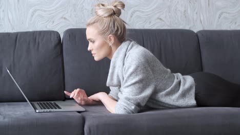 Happy-blond-woman-lying-prone-on-sofa-and-working-on-laptop-computer