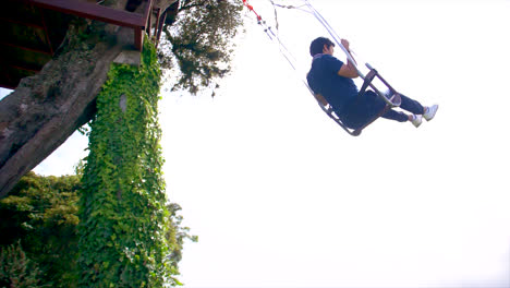 Young-man-goes-high-on-rope-swing-by-treehouse-in-Ecuador,-side-slomo