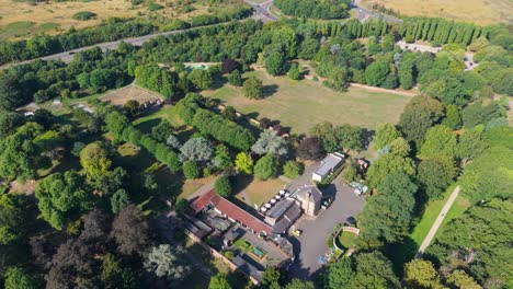 Wide drone orbit showing Cedar’s Park main buildings in Cheshunt, England, with lush green summer trees, surrounding parkland, maze, and clear architectural details