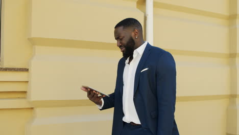 Upset-businessman-walking-with-gadget-outdoors.-Afro-guy-using-tablet-outside