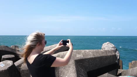 Mujer-Joven-Tomando-Fotos-En-Su-Celular-Del-Océano-En-Un-Muelle-Con-El-Viento-Soplando-Su-Cabello-En-Un-Día-Soleado