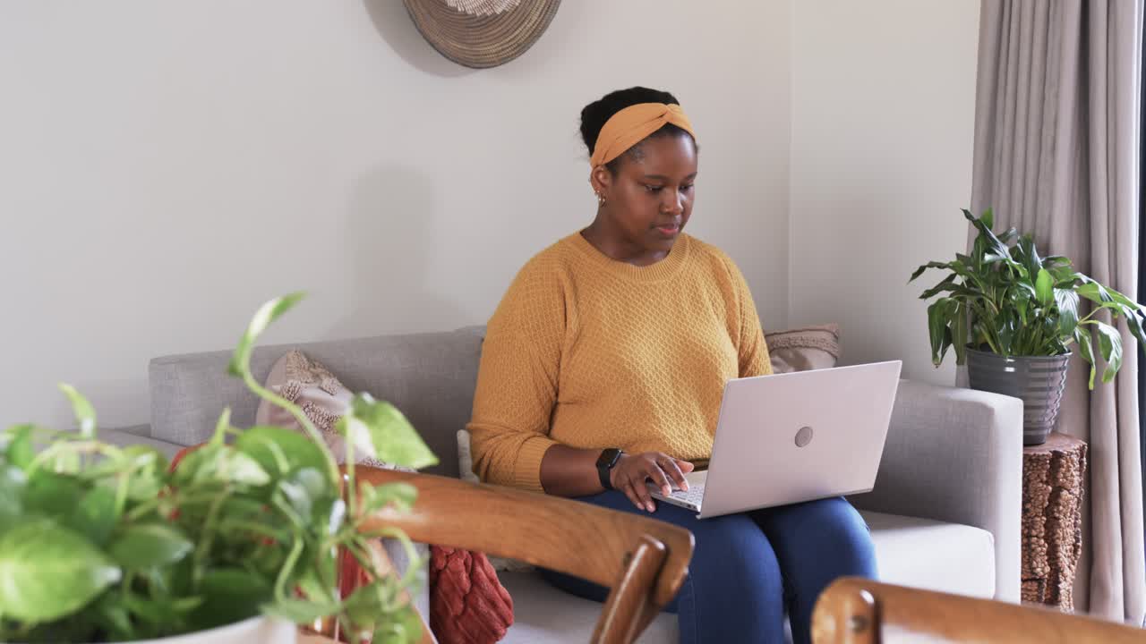 Entering home African American woman sitting on couch, typing on laptop, pausing and resuming work