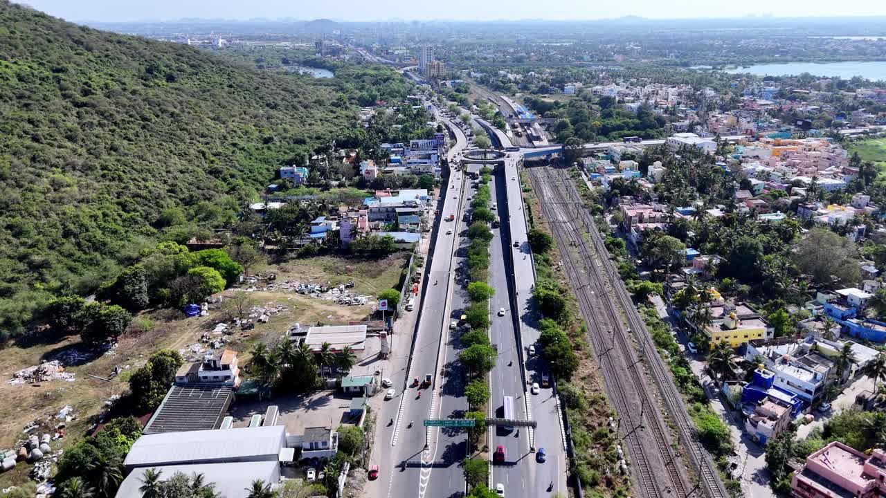 Aerial view of a major highway Grand Southern Trunk Road (GST Road) and railway tracks in Chennai, flanked by a green hill on one side and urban development on the other, under a clear sky.
