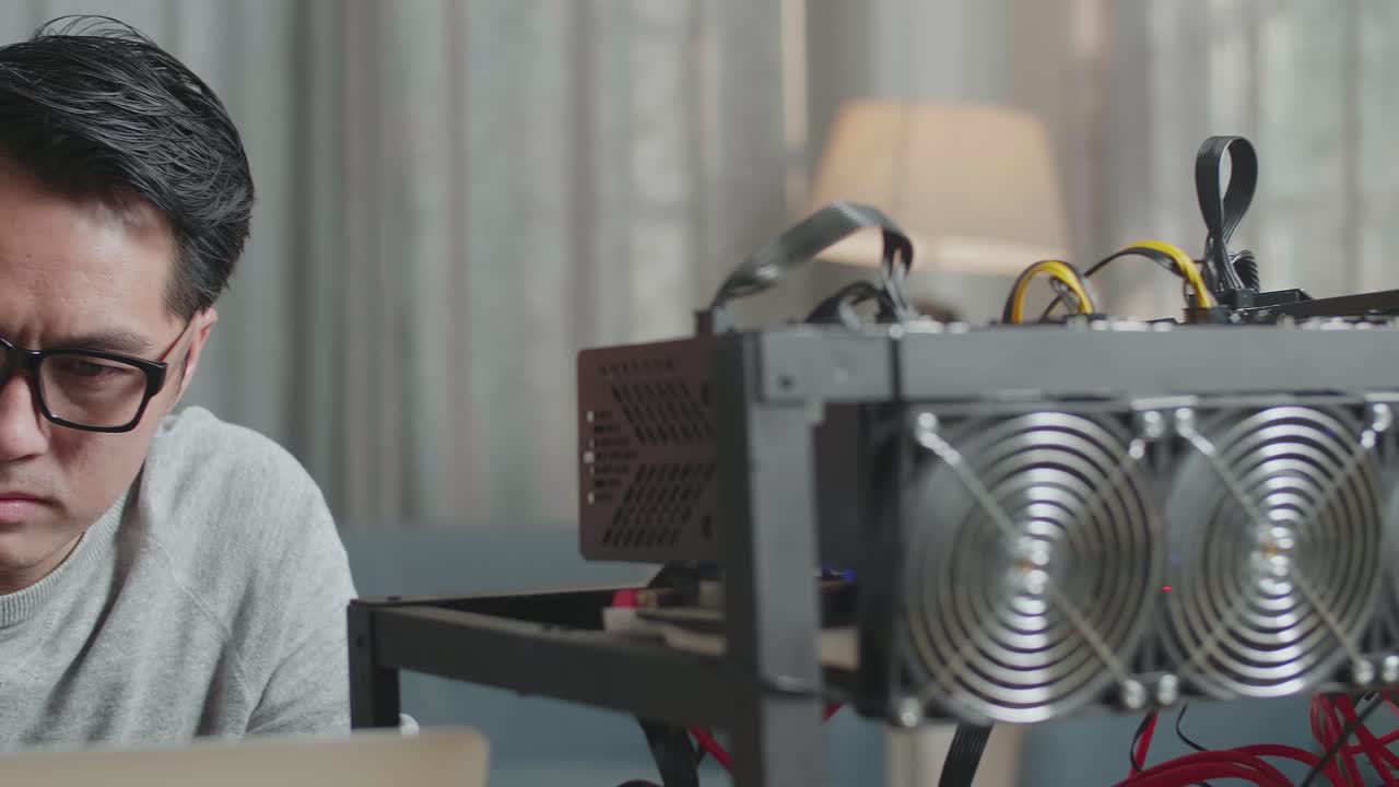 심각한 아시아 남자가 안경을 쓰고 노트북과 채굴 기계를 사용하여 암호화폐를 채굴합니다.