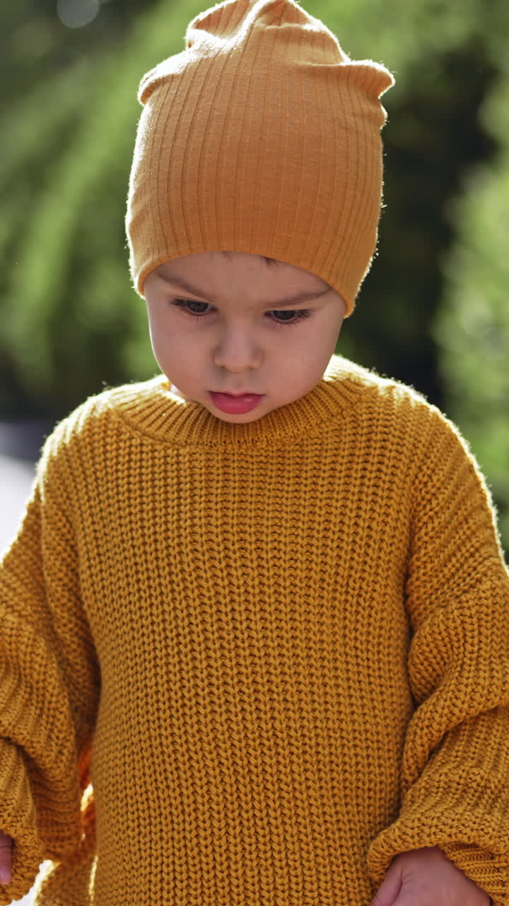 Caucasian toddler wearing yellow cap and knitted sweater. Baby boy is outdoors on sunny weather. Blurred backdrop. Vertical video