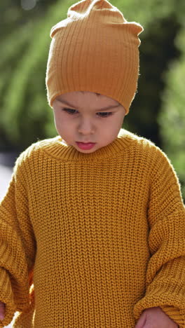 caucasian toddler wearing yellow cap and knitted sweater. baby boy is outdoors on sunny weather. blurred backdrop. vertical video