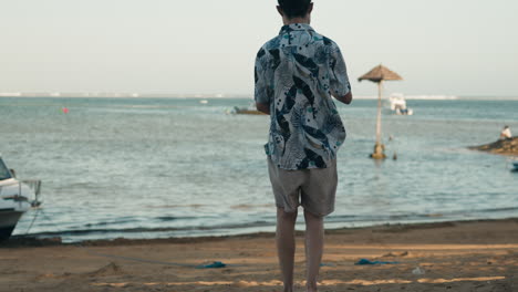 Un-Hombre-Asiático-Camina-Hacia-La-Playa-De-Bali-De-Vacaciones-Con-Gafas-De-Sol-Sosteniendo-El-Teléfono
