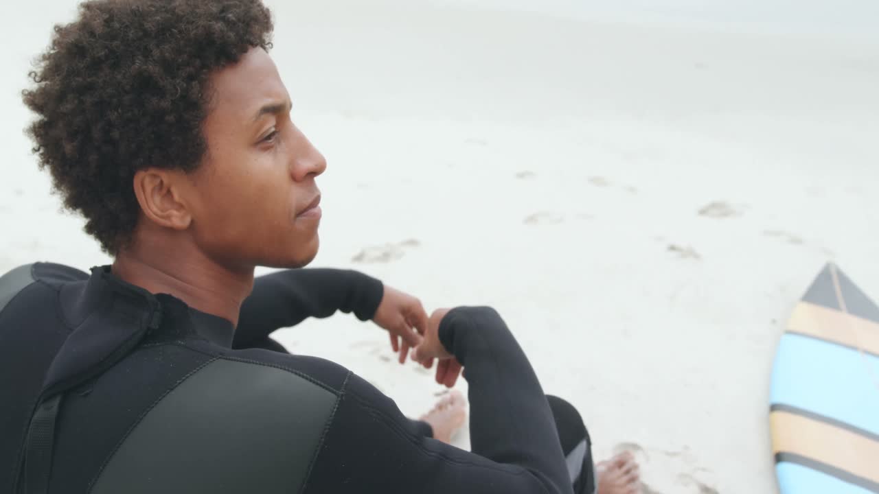 Side view of African American male surfer relaxing with surfboard on the beach 4k