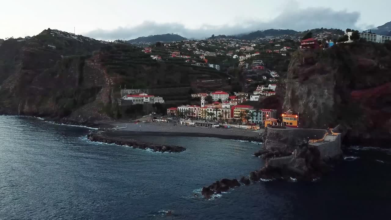 Ponta do Sol, Madeira, durante una sera nuvolosa, che mostra le case colorate che coprono le ripide scogliere, la spiaggia di sabbia nera e la costa rocciosa, ripresa aerea introduttiva con drone