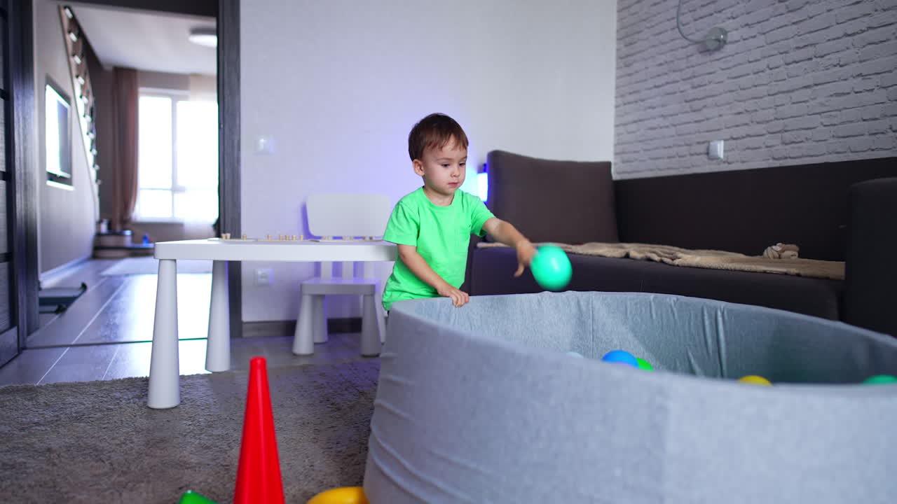 Eighteen months old toddler plays in the room with a dry pool. Baby boy throws a ball into the basin.