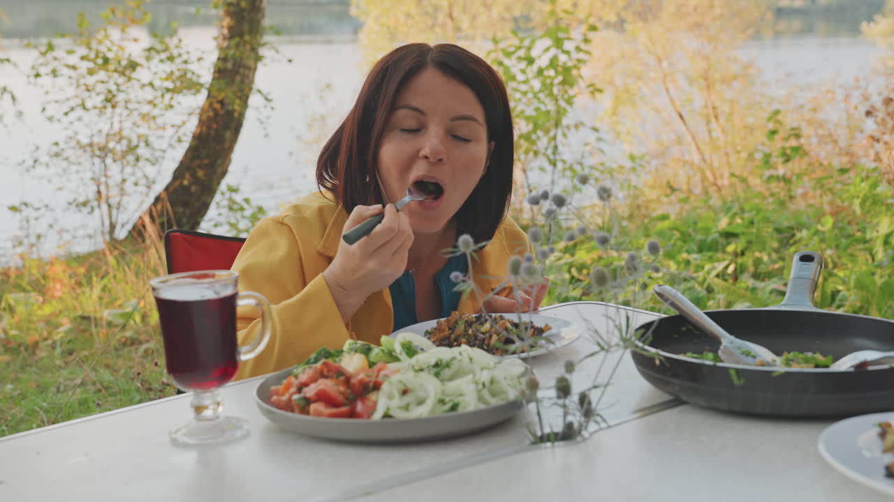 Mujer caucásica acampando junto al lago, probando una comida, bloguera de viajes degustando una ensalada y platos a la sartén, bebida roja en copa de tallo, telón de fondo costero con juncos y árboles, ambiente reflexivo, brisa suave, informal