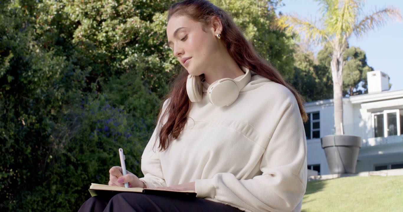 Woman settling on grassy slope in yard, writing in notebook with pen and headphones, journaling