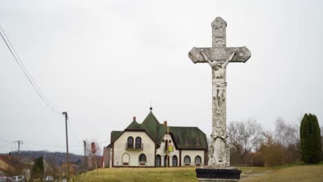 a stone crucifix with jesus in the foreground, with a historic building and village scenery in the background