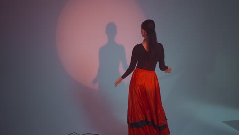 a lady in a black and red gown dances facing the wall, moving her body and clapping hands slowly in the air against a red and grey background