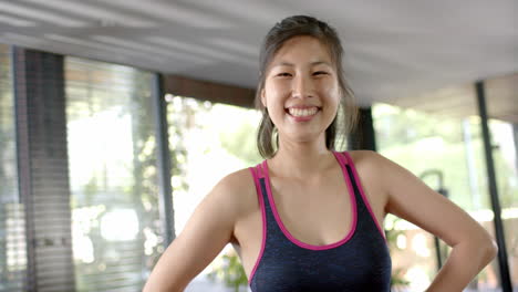 happy asian woman with arms akimbo looking at camera and smiling in sunny home gym, slow motion