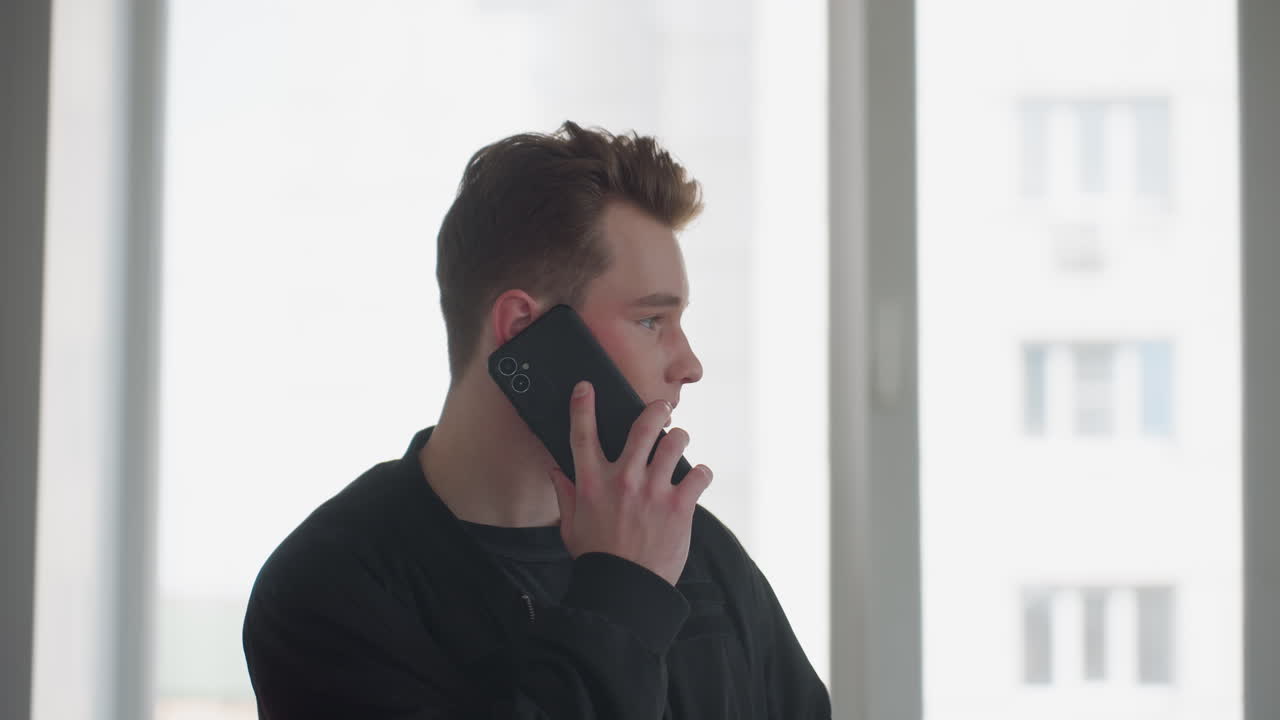 Young boy in black jacket holds smartphone to ear while speaking, standing near large window with cityscape blurred outside, soft natural light illuminating contemplative expression