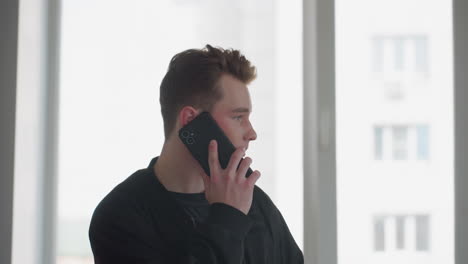 young boy in black jacket holds smartphone to ear while speaking, standing near large window with cityscape blurred outside, soft natural light illuminating contemplative expression
