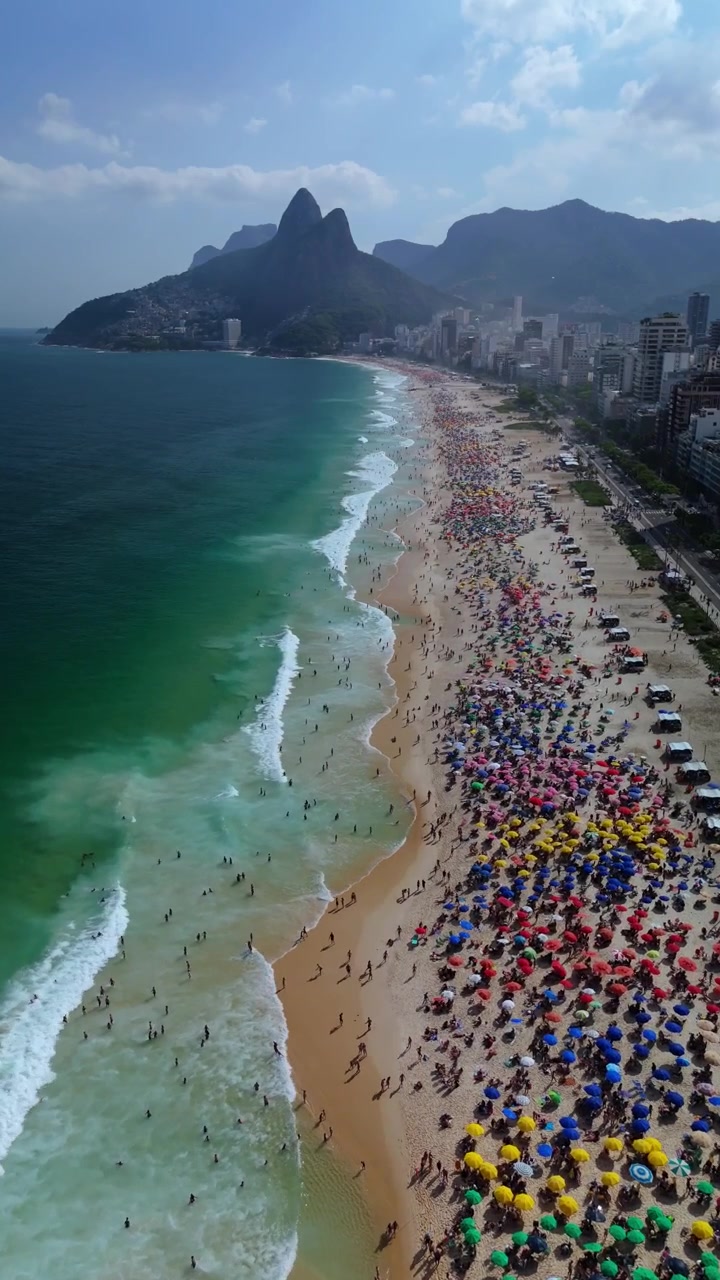 Drone vertikal terbang di atas Pantai Ipanema yang ramai, menunjukkan keramaian, ombak, garis pantai, dan cakrawala Rio de Janeiro.