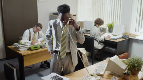 Afroamerikanischer-Geschäftsmann,-Der-Neben-Seinem-Schreibtisch-Arbeitet-Und-In-Einem-Vintage-Büro-Telefoniert.