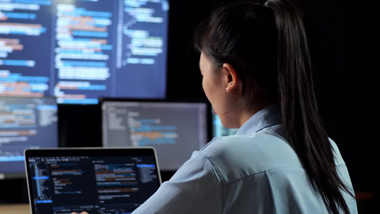 vista de atrás de una programadora asiática estirándose mientras escribe código en una computadora portátil usando múltiples monitores que muestran la base de datos en los escritorios de ventana de terminal en la oficina