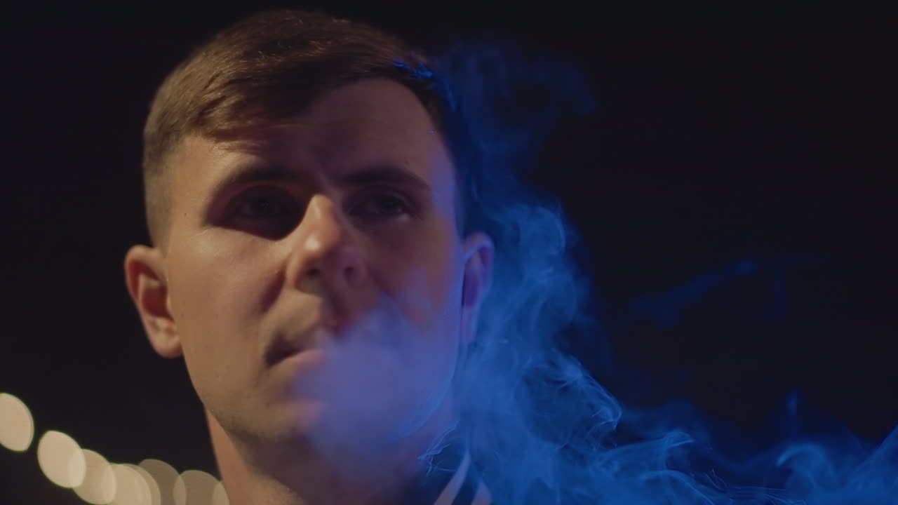 빽빽한 푸른색 구름을 내뿜는 백인 남성, 네온 조명이 켜진 거리에서 극적인 클로즈업, 얼굴을 뒤덮는 증기, 그림자가 드리워진 이목구비, 반사적인 표정, 영화 같은 긴장감, 도시의 안개와 맥동하는 조명