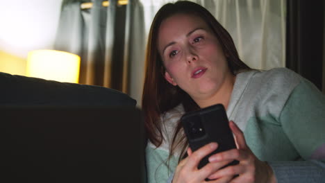 Close-Up-Of-Woman-Sitting-On-Sofa-At-Home-At-Night-Streaming-Or-Watching-Movie-Or-Show-On-Laptop-And-Scrolling-Internet-On-Phone-1