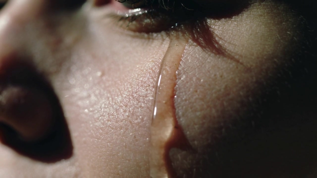 Holding still, female face showing tear forming at lower eyelid and running down cheek, close-up