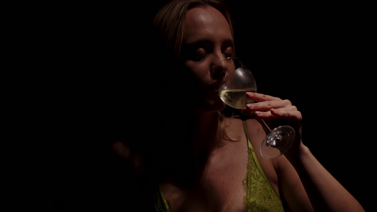Primo piano cinematografico di una donna attraente che beve un bicchiere di vino bianco in una stanza buia al rallentatore.