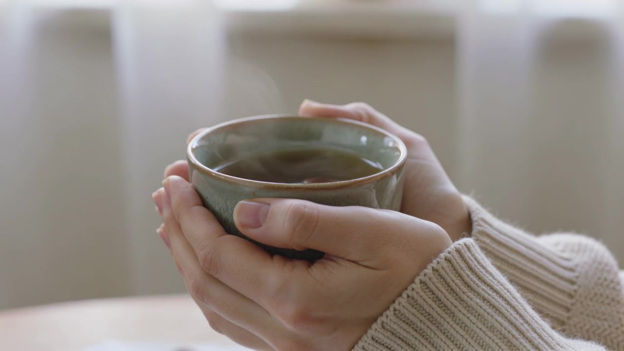 Mani adulte con polsini in maglia color crema che si scaldano al vapore vicino alla finestra, tazza grigio-verde che emette vapore