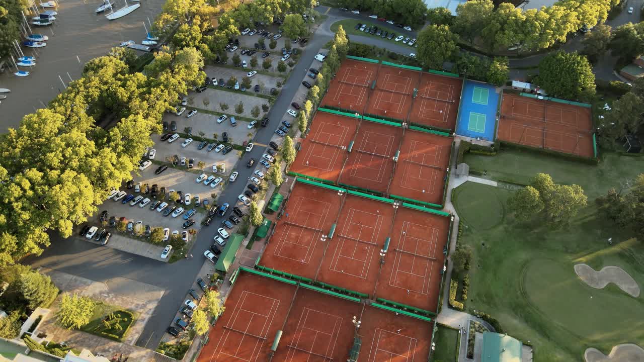 Ripresa aerea orbitante su campi da tennis in terra battuta, un campo da golf e un parcheggio in un club sportivo privato all'ora d'oro