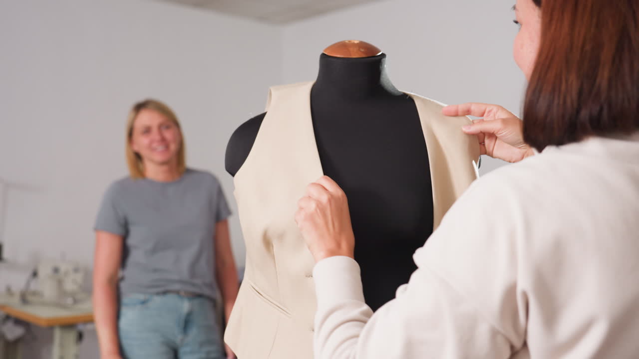 Tailor puts on yellow blazer onto black dress form with precision while partner admires fit and gives approval in bright sewing studio filled with samples and tools in modern workshop
