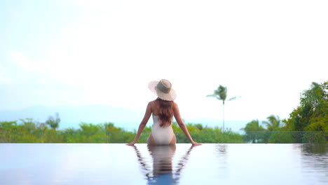 a healthy, fit, sexy woman sits on the edge of a resort infinity pool with her back to the camera as she looks out over the ocean