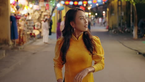 Young-Vietnamese-woman-in-traditional-dress-walks-down-night-street-in-Hoi-An,-Vietnam-in-slow-motion