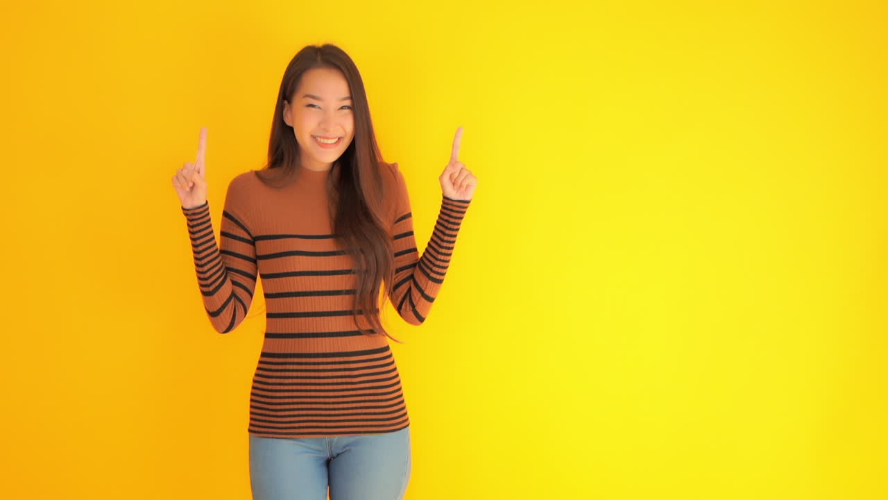 Smiling Asian girl holding up both fingers pointing upwards. Copy Space Yellow Background