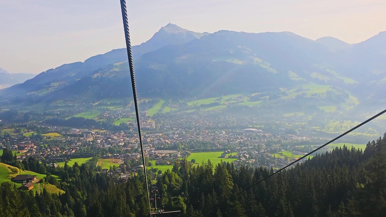 Una cabinovia che sale sopra un villaggio di montagna, con cabine rosse, case chalet, pascoli verdi, una fitta pineta e aspre creste alpine in un clima estivo luminoso per il viaggio.